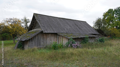 An autumn cloudy day. An old barn. Behind the barn you can see an abandoned house with boarded up windows.