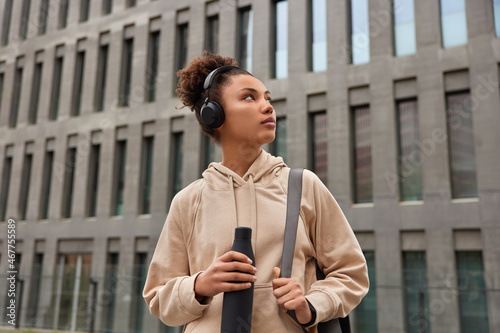 Outdoor shot of sportswoman prepares for workout carries karemat necessary for training holds bottle of water to hydrate herselt listens music in headphones concentrated on modern city building photo