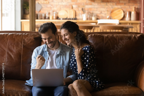 Excited winners. Overjoyed married couple sit on large sofa at modern cottage interior use laptop app laugh win lottery prize. Amazed young spouses raise fists yes get great offer for family business