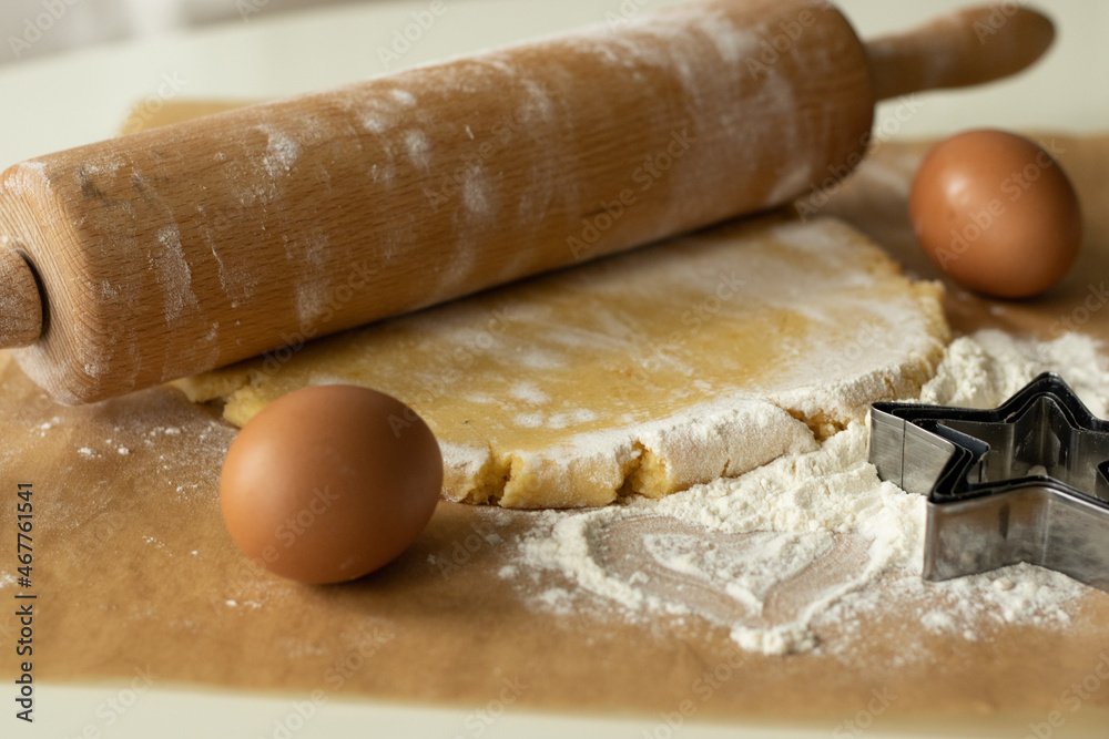 The dough, eggs and flour prepared for cooking lie on parchment paper, next to it lies a wooden rolling pin and an asterisk shape for baking. The dough, eggs and flour prepared for cooking lie on parchment paper, next to it lies a wooden rolling pin and an asterisk shape for baking.
