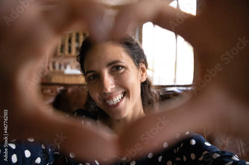 I love you. Portrait of smiling young woman look at digital camera through heart shape made of united fingers. Happy millennial latin lady feeling love talk by video call to boyfriend in romantic mood