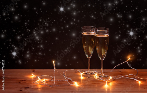 Glasses with champagne and glowing christmas lights on dark background. Selective focus.