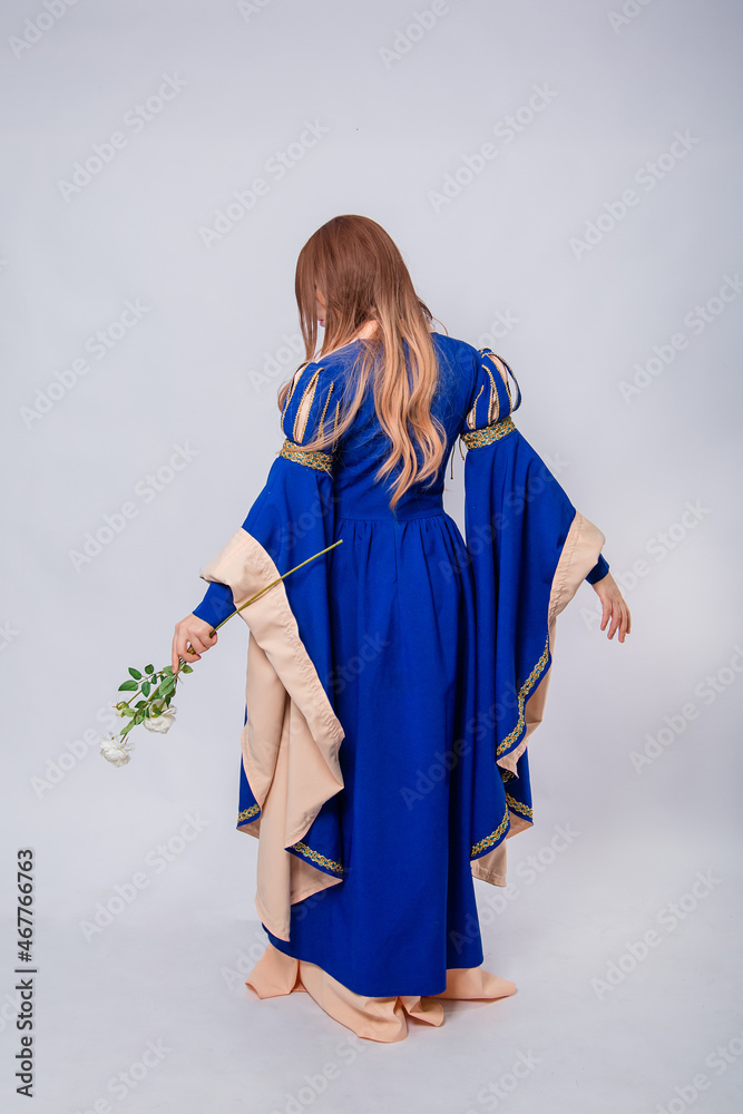 Fototapeta premium Full-length portrait, back view, attractive woman in medieval, fantasy blue-beige dress with long, flared sleeves posing with a white rose in her hands, isolated on white background.
