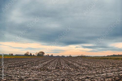 Freshly ploughed field of heavy clay under a grey sky, just before sunset. Trees and farms in the background.
 photo