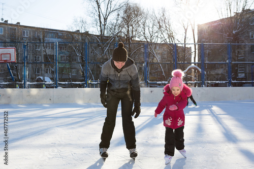 Dad teaches his little daughter to ice skate on a skating rink in the courtyard of multi-storey buildings in the city. Frosty winter sunny day, active winter sports and lifestyle