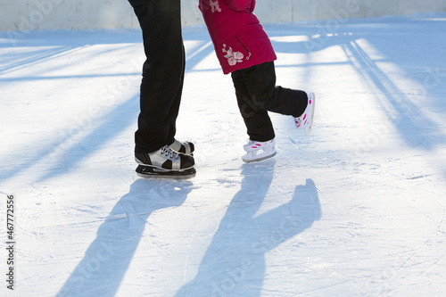 Dad teaches his little daughter to ice skate on a skating rink in the courtyard of multi-storey buildings in the city. Frosty winter sunny day, active winter sports and lifestyle