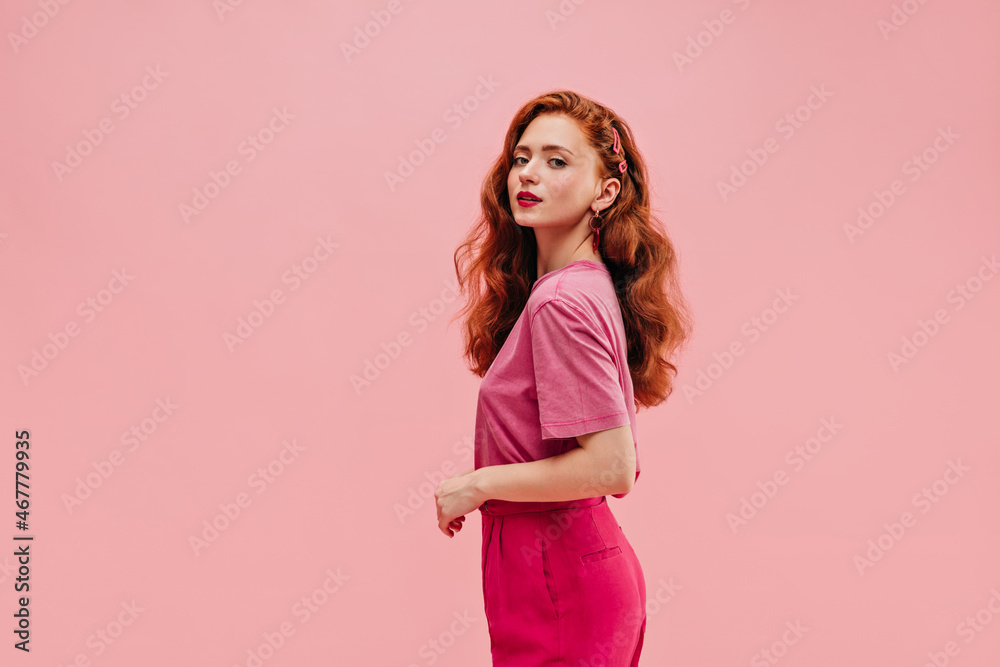 Cute caucasian young girl with fluffy wavy hair on one side on pink background with place for text. Red-haired lady in wide T-shirt and crimson pants looks at camera with her mouth open.
