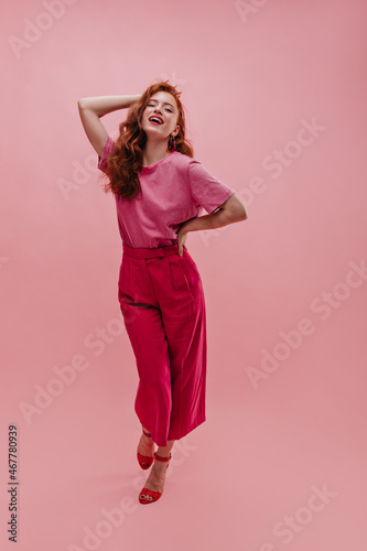 Full length fashionable caucasian young lady posing walking forward holding her waist and hair from behind in pink room. Tipping her head red-haired girl with open mouth in T-shirt, pants, shoes.