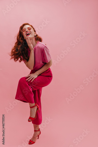 Full length fashionable caucasian young girl laughs hard with mouth wide open on isolated background. Red-haired lady with sunglasses on face squeezes her leg to knee, dressed in casual pink clothes.