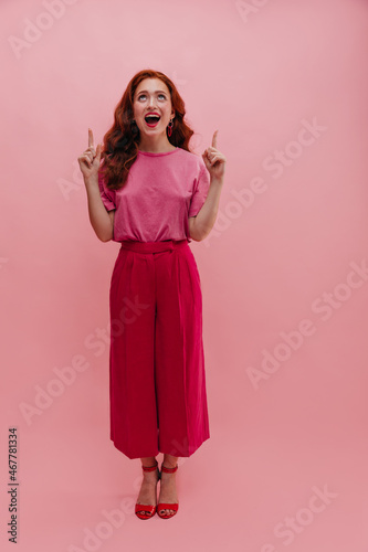 Full length pretty slim caucasian young woman pointing her fingers up and looking at same indoors. Redhead teenager stands straight with open mouth, dressed in casual pink clothes.