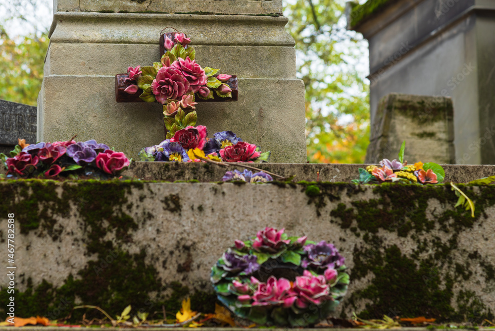 Cemetery in autumn . Grief and memorial. Flower decorated ceramic cross and wreaths over tomb. Mourning vintage background. Funeral services retro design concept. Selective focus on cross.