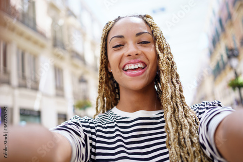 Black woman with afro braids taking a smiling selfie in an urban street with a smartphone.