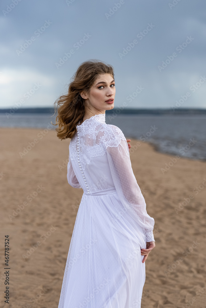 Happy young girl in a white dress on the river bank. Stormy sky over the river