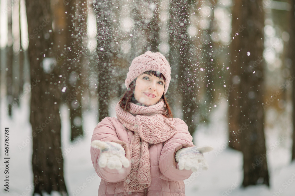 Hello winter. Smiling happy young woman in pink coat, cap and scarf, outdoors in the forest in winter, throwing up snow and having fun