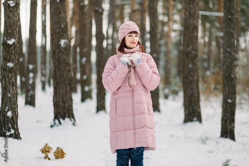 Outdoor winter portrait of pretty young woman wearing pink coat and stylish knitted hat and scarf, standing in beautiful snowy forest in front of pine trees and enjoying walk on frosty day