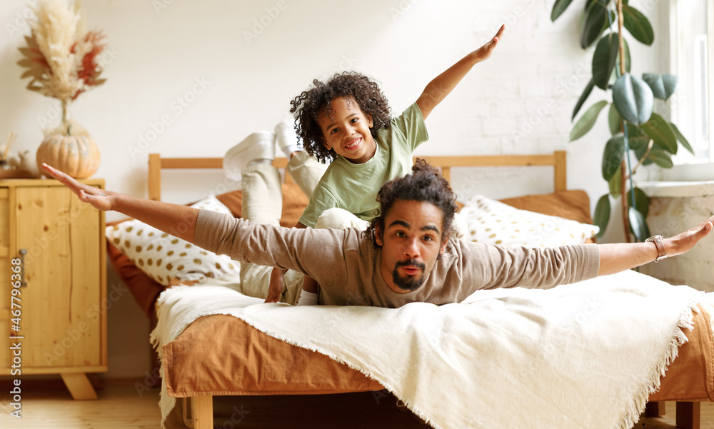 Happy african american family father and son in flying pose lying on bed at home - obrazy, fototapety, plakaty 