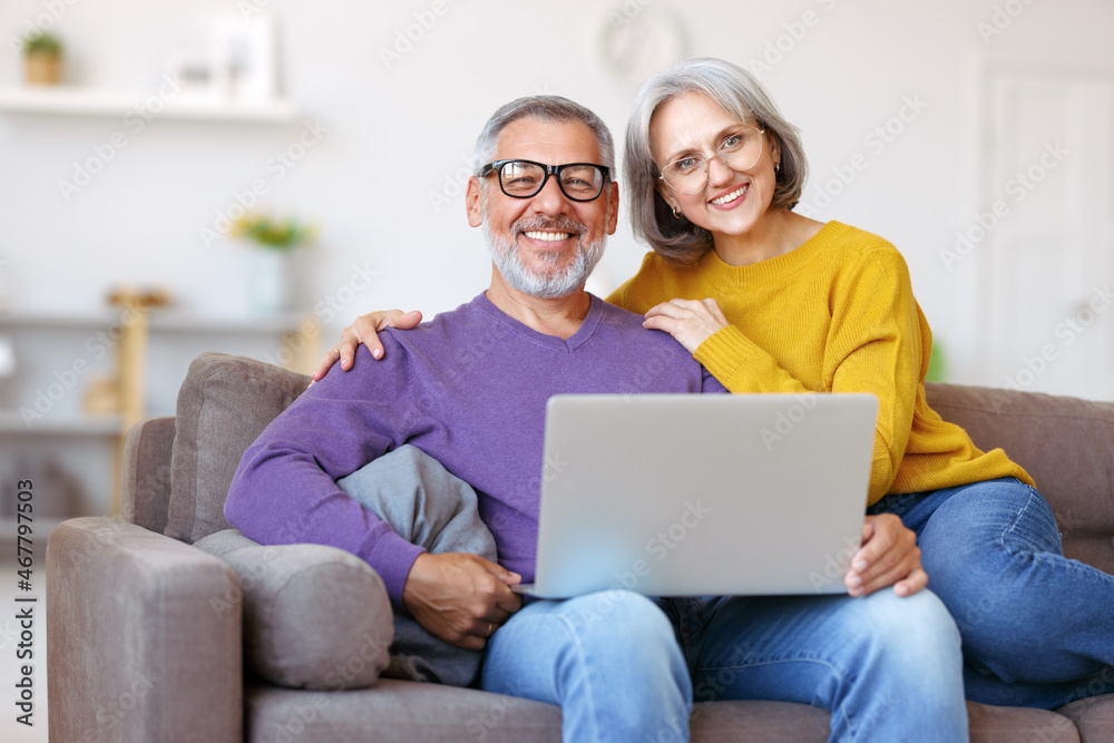 Beautiful mature family couple looking at laptop with smile on face while spending time together Beautiful mature family couple looking at laptop with smile on face while spending time together