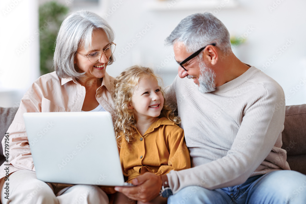 Smiling senior grandparents and cute little girl granddaughter watching cartoons on laptop