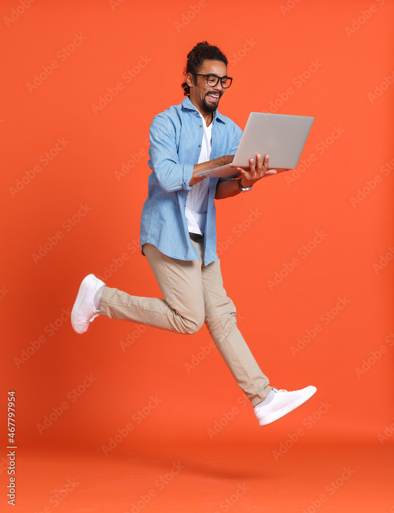 Young happy overjoyed african american man with laptop computer jumping over orange background