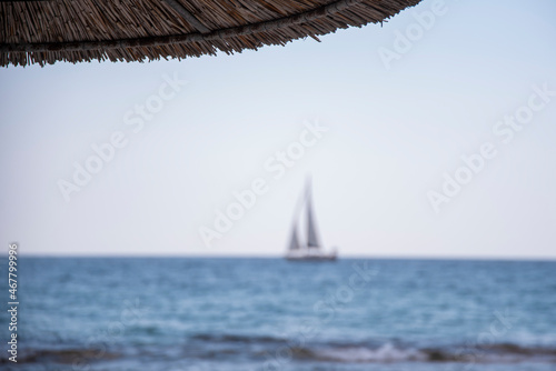 Reed umbrella. Silhouette of beautiful modern sailboat sailing blue Aegean sea. Summer adventure, active vacation in Nea Skioni, Greece.