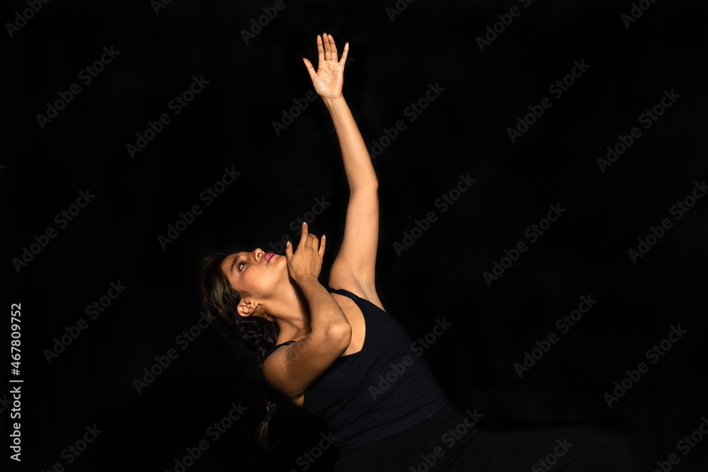 Hispanic young woman performing body movements. Woman raises arms and looks up dramatically isolated on black background