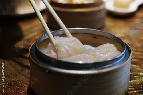 A close-up picture of bamboo chopsticks picking up white white Chinese stuffed steamed dumpling, also known as dim sum from a round bamboo basket on a nice dinner table.
