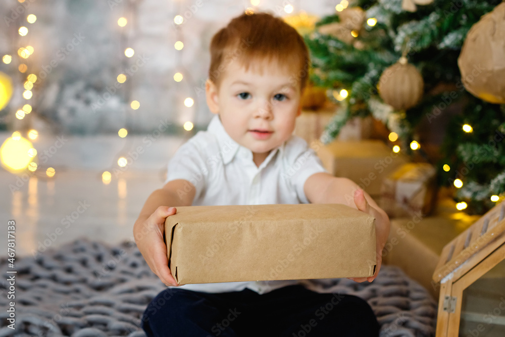 Selective focus. A boy is sitting on the floor with a gift box for a room decorated for Christmas against the background of lights of garlands. He hands the gift to the camera. Selective focus. A boy is sitting on the floor with a gift box for a room decorated for Christmas against the background of lights of garlands. He hands the gift to the camera.