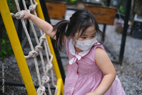 A cute young Asian girl, with white face mask, is playing alone on a jungle gym in a playground, climbing a ramp and nets made from rope during Covid-19 pandemic.