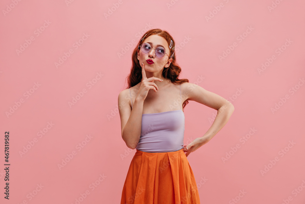 Pensive caucasian young girl looks to side pursing her lips on pink background. Redhead lady holds her chin with finger, wearing lilac top and orange skirt. Thoughtfulness concept Pensive caucasian young girl looks to side pursing her lips on pink background. Redhead lady holds her chin with finger, wearing lilac top and orange skirt. Thoughtfulness concept