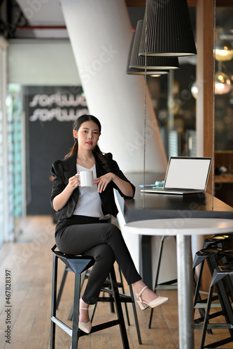 Elegant Asian young businesswoman sits on the chair and hold coffee cup in a modern office.