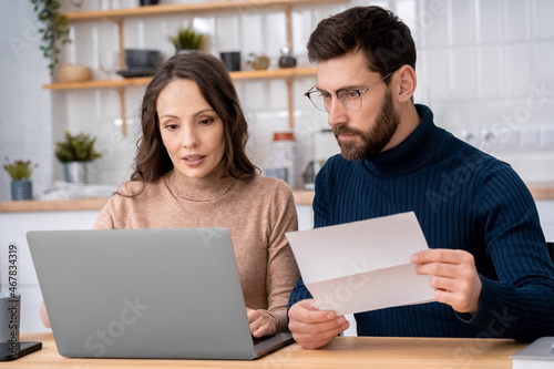 Family married couple looking at computer pc screen making the utility payments sitting at home kitchen. Wife holding paper bills and husband paying online using banking application on laptop indoors photo