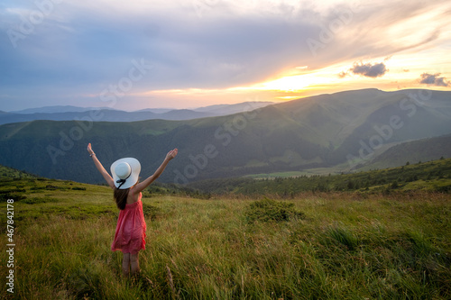 Back view of young happy woman traveler in red dress standing on grassy hillside on a windy evening in summer mountains with outstretched arms enjoying view of nature at sunset