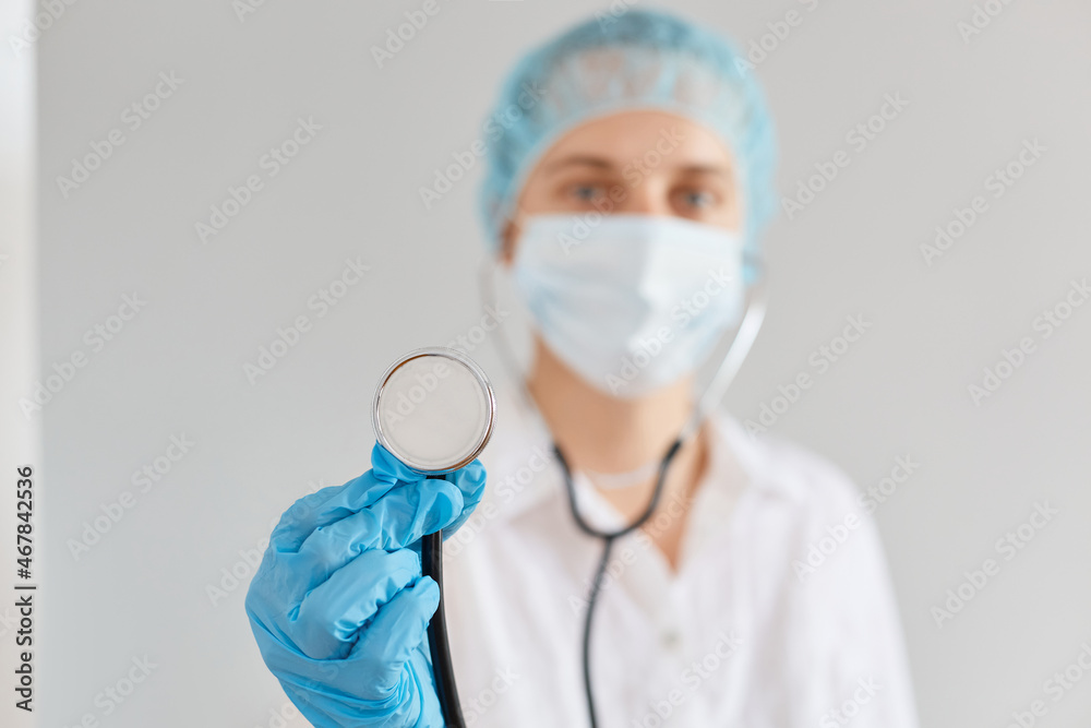 Indoor blurred shot of young adult woman doctor wearing medical cap, gloves and protective mask showing stethoscope to camera, equipment for diagnostic.