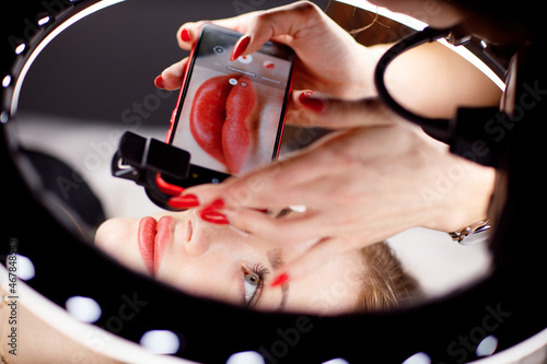 Cosmetologist making photo of work on mobile phone red permanent make up tattoo on young woman lips.