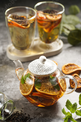 A transparent tea pot filled with black tea, oranges and mint on a grey surface