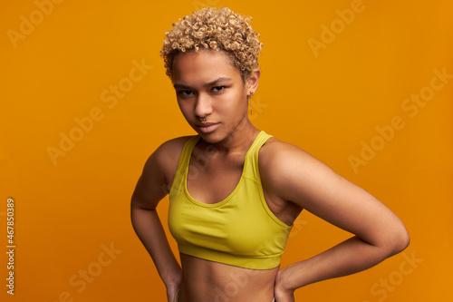 Disappointed young dark-skinned woman with nose ring and blonde Afro curls looking at camera with discontent, angry face expression isolated on orange wall holding hands in bossy position on waist