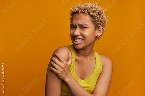 Portrait of pretty dark-skinned girl of 20s feeling pain in upper arm, tighten her teeth, trying to get rid of feeling squeezing shoulder. Body ache, trauma, muscle pain. Human emotions