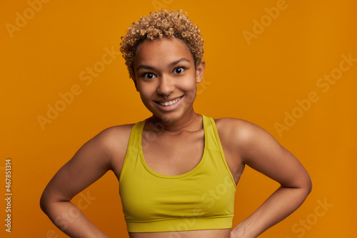 Attractive young athletic female with small blonde poodles and nose piercing posing against orange studio wall with surprised face expression, looking at camera with amazed smile, feeling astonished