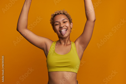 Funny cropped picture of cheerful carefree young blonde woman of mixed race holding hands high, sticking tongue, fooling around making faces, having cute short poodles, wearing yellow stylish top photo
