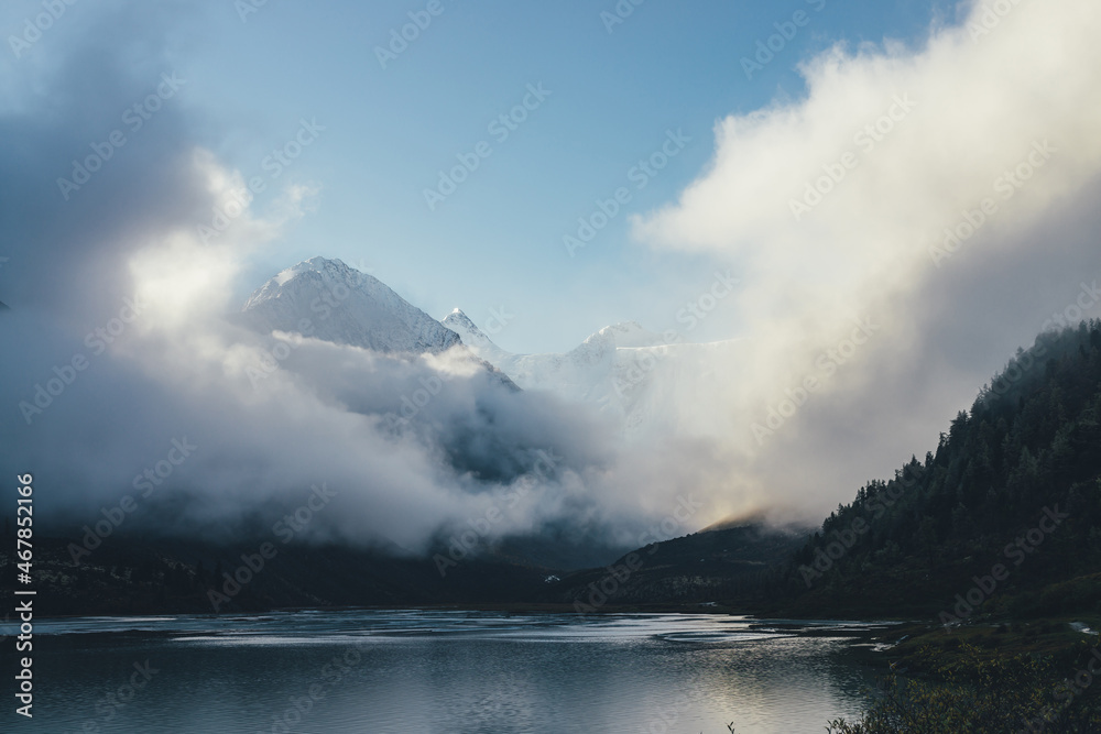 Beautiful view of snow-capped mountains above thick clouds in sunshine. Scenic mountain landscape with white-snow peak among dense low clouds in blue sky. Wonderful alpine scenery with snowy pinnacle.
