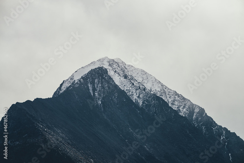 Dark atmospheric landscape with high black rocky mountain top with snow in cloudy sky. Dramatic mountain landscape with snow peaked top in haze in overcast weather. Gloomy view to awesome rocks in fog