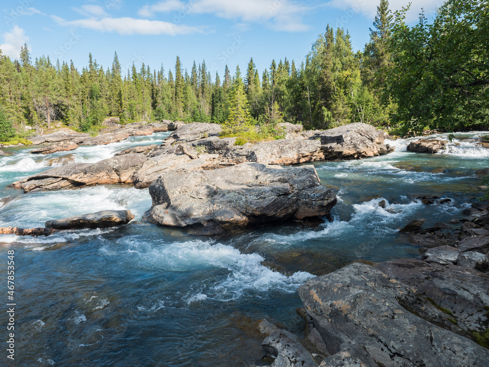 Beautiful northern landscape with rapids and cascades of wild river Kamajokk, boulders and spruce tree forest and hills in Kvikkjokk village in Swedish Lapland. Summer sunny day, blue sky white clouds Beautiful northern landscape with rapids and cascades of wild river Kamajokk, boulders and spruce tree forest and hills in Kvikkjokk village in Swedish Lapland. Summer sunny day, blue sky white clouds