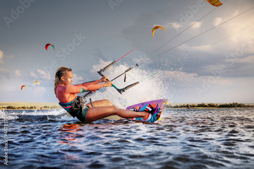 Professional kitesurfer young caucasian woman glides on a board along the sea surface at sunset against the backdrop of beautiful clouds and other kites. Active water sports