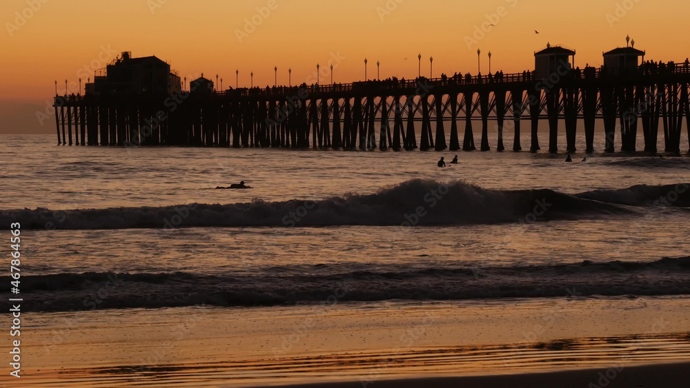 Obraz premium Wooden pier silhouette at sunset, California USA, Oceanside. Waterfront surfing resort, pacific ocean tropical beach. Summertime sea coastline vacations atmosphere. Surfers waiting for wave in water.