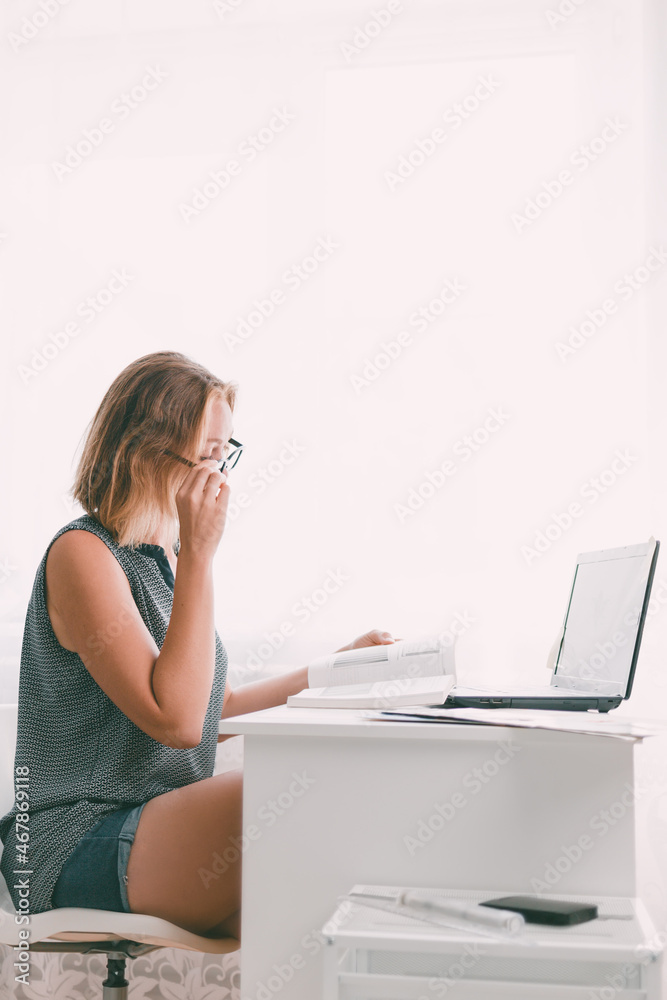 Young woman with glasses sitting at the table and leafing through a book. Nearby are a laptop and office supplies. Interior is white. Copy space. Vertical orietation Young woman with glasses sitting at the table and leafing through a book. Nearby are a laptop and office supplies. Interior is white. Copy space. Vertical orietation