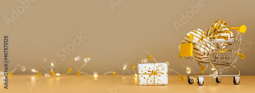 Online christmas shopping and sale. Shopping cart with gift boxes on golden background with lights. Supermarket trolley full presents. Banner with copy space