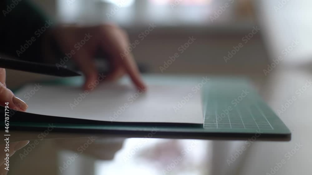 Close-up side view of hands of female designer putting white paper on green rubber cutting mat and makes mark at desk on blurred background of window, selective focus. Shooting in slow motion. Close-up side view of hands of female designer putting white paper on green rubber cutting mat and makes mark at desk on blurred background of window, selective focus. Shooting in slow motion.