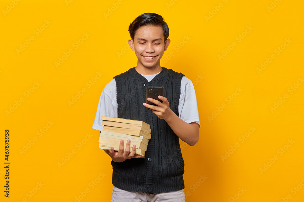 Happy Asian young man looking at smartphone screen and holding book on yellow background Happy Asian young man looking at smartphone screen and holding book on yellow background