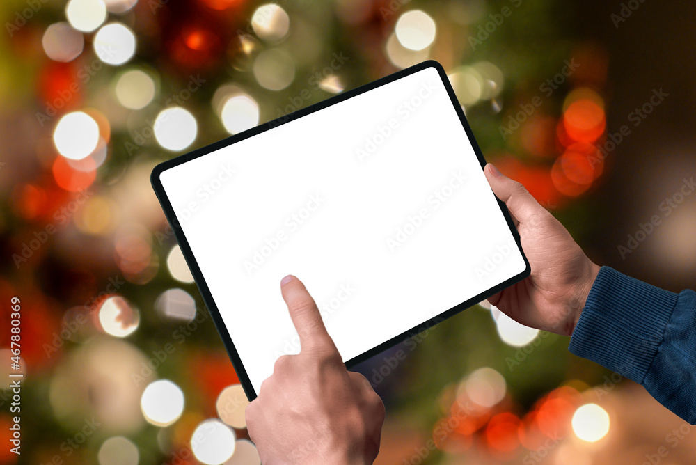 Tablet in the hands with the lights of the Christmas tree behind. Isolated display for design promotion. Season online shopping concept Tablet in the hands with the lights of the Christmas tree behind. Isolated display for design promotion. Season online shopping concept
