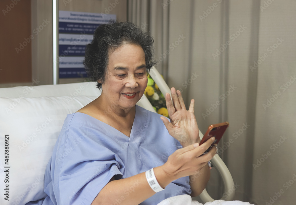 cheerful Asian senior female patient sitting in hospital bed, smiling and looking at mobile phone, video calling and waving to her family. Elderly health concept. cheerful Asian senior female patient sitting in hospital bed, smiling and looking at mobile phone, video calling and waving to her family. Elderly health concept.